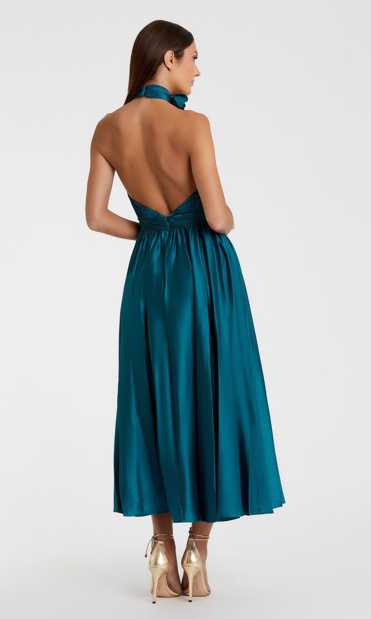 A woman in the Mac Duggal 49674 ocean blue halter midi wedding guest dress with an open back and gold high heels stands with her back to the camera against a plain background.